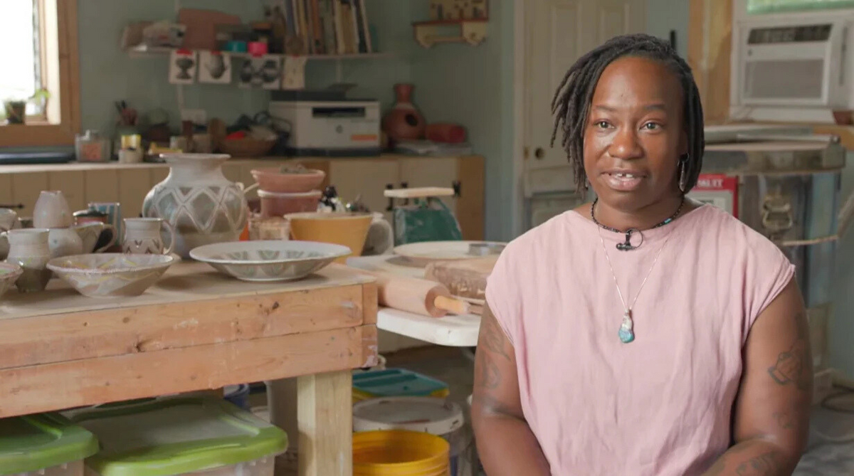 Load video: A video segment with Osa Atoe about how she draws inspiration from the Florida landscape. For the interview, she is seated in her studio surrounded by finished pottery pieces as well as her tools and equipment, include an electric kiln in the background.